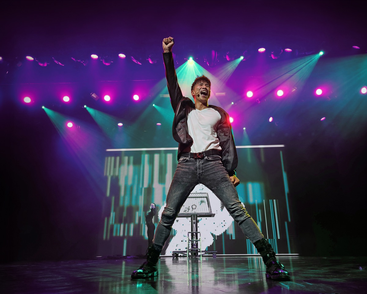 A performer on stage with one fist raised, under purple and green lights